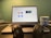 A laptop displaying a video conferencing application screen is placed on a desk. In front of the laptop, there is a pair of black headphones resting on the desk. To the right, a blue mug with 'Digital Startup Challenge' printed on it is positioned. The background wall is green, creating a subtle contrast with the objects in the foreground.