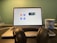 A laptop displaying a video conferencing application screen is placed on a desk. In front of the laptop, there is a pair of black headphones resting on the desk. To the right, a blue mug with 'Digital Startup Challenge' printed on it is positioned. The background wall is green, creating a subtle contrast with the objects in the foreground.