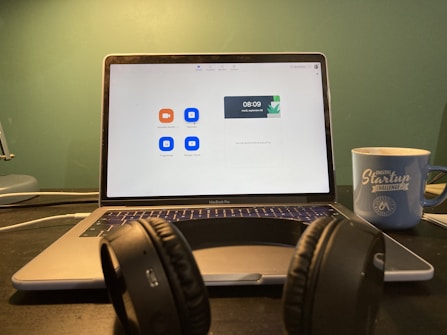 A laptop displaying a video conferencing application screen is placed on a desk. In front of the laptop, there is a pair of black headphones resting on the desk. To the right, a blue mug with 'Digital Startup Challenge' printed on it is positioned. The background wall is green, creating a subtle contrast with the objects in the foreground.