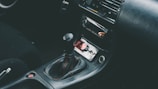 The interior of a car showcasing a manual gear shift in the center console area, surrounded by a dark leather upholstery. The dashboard includes a radio and air conditioning controls. A red button and a small keychain are visible in the storage compartment.