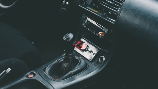 The interior of a car showcasing a manual gear shift in the center console area, surrounded by a dark leather upholstery. The dashboard includes a radio and air conditioning controls. A red button and a small keychain are visible in the storage compartment.