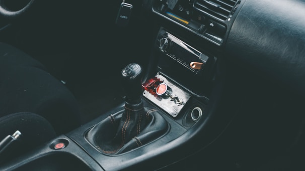 The interior of a car showcasing a manual gear shift in the center console area, surrounded by a dark leather upholstery. The dashboard includes a radio and air conditioning controls. A red button and a small keychain are visible in the storage compartment.