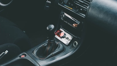 The interior of a car showcasing a manual gear shift in the center console area, surrounded by a dark leather upholstery. The dashboard includes a radio and air conditioning controls. A red button and a small keychain are visible in the storage compartment.