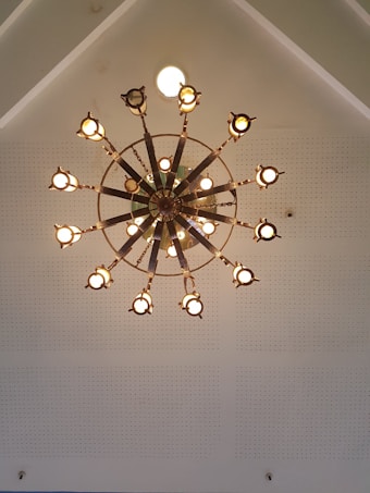 An ornate, circular chandelier with multiple lamps evenly spaced along its radial arms hangs from a ceiling. The ceiling is white and features perforated acoustic tiles. The light from the chandelier illuminates the space with a warm glow.