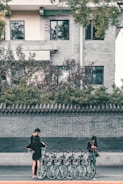 A row of bicycles is lined up on the sidewalk in front of a brick wall. Two people, both wearing black clothing, are interacting with the bicycles, possibly checking or renting them. One person is standing slightly apart, focused on a mobile device. The scene is set against a residential building featuring windows and a tile-roofed wall with greenery on top.