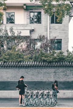 A row of bicycles is lined up on the sidewalk in front of a brick wall. Two people, both wearing black clothing, are interacting with the bicycles, possibly checking or renting them. One person is standing slightly apart, focused on a mobile device. The scene is set against a residential building featuring windows and a tile-roofed wall with greenery on top.