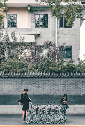 A row of bicycles is lined up on the sidewalk in front of a brick wall. Two people, both wearing black clothing, are interacting with the bicycles, possibly checking or renting them. One person is standing slightly apart, focused on a mobile device. The scene is set against a residential building featuring windows and a tile-roofed wall with greenery on top.