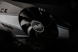 A close-up view of a computer graphics card featuring a large fan with the ASUS logo at the center. The sleek design emphasizes the metallic and black components, highlighting the hardware's modern technology.