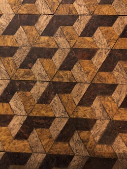 An intricate geometric pattern featuring a series of interlocking triangles and parallelograms. The design uses a textured, wood-like material with a combination of brown, tan, and ochre shades, giving it a rustic appearance.