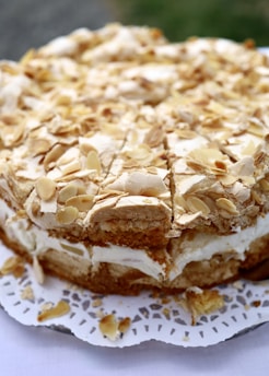 A close-up of a cake topped with almond slices. The cake features layers of cream and crust, with a lacy doily underneath. The texture appears light and fluffy, with the almonds adding a crunchy contrast.