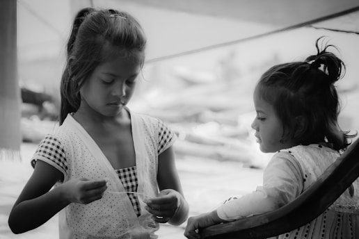 Two young girls are sitting together in a calm setting. One girl is focused on a task in her hands, while the other looks on from a nearby chair. The scene is captured in black and white, adding a timeless and contemplative feel.