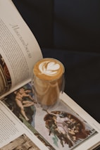 A glass with a layered latte featuring artful foam on top, along with an open book displaying Renaissance art. The book's text is in a foreign language, showcasing classic paintings with rich detail.