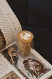 A glass with a layered latte featuring artful foam on top, along with an open book displaying Renaissance art. The book's text is in a foreign language, showcasing classic paintings with rich detail.