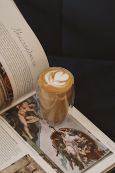A glass with a layered latte featuring artful foam on top, along with an open book displaying Renaissance art. The book's text is in a foreign language, showcasing classic paintings with rich detail.