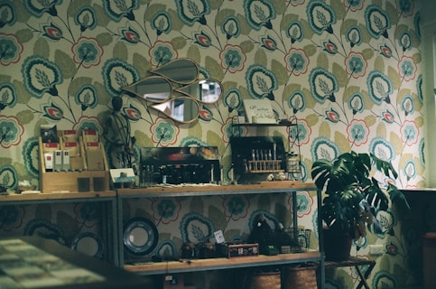 A cozy interior with vintage-style wallpaper featuring a floral pattern as a backdrop. Shelves hold a variety of items including small art pieces, a decorative mirror, books, and potted plants. The decor creates a rustic and inviting atmosphere.
