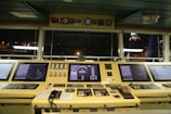 A ship's bridge control panel containing multiple electronic displays, gauges, and communication devices. The displays show navigational radar and mapping information. The panel is well-lit, with a view of the ship's surroundings visible through large windows, indicating nighttime.