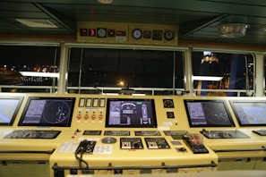 A ship's bridge control panel containing multiple electronic displays, gauges, and communication devices. The displays show navigational radar and mapping information. The panel is well-lit, with a view of the ship's surroundings visible through large windows, indicating nighttime.