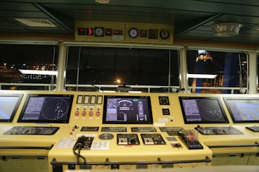 A ship's bridge control panel containing multiple electronic displays, gauges, and communication devices. The displays show navigational radar and mapping information. The panel is well-lit, with a view of the ship's surroundings visible through large windows, indicating nighttime.
