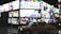 Brightly lit billboards display colorful advertisements featuring various people and text in an urban nighttime setting. A taxi is parked on the wet street while several pedestrians holding umbrellas walk by.