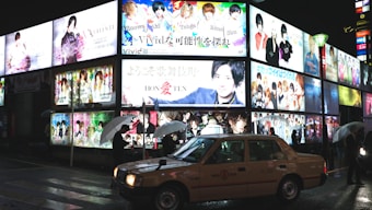 Brightly lit billboards display colorful advertisements featuring various people and text in an urban nighttime setting. A taxi is parked on the wet street while several pedestrians holding umbrellas walk by.
