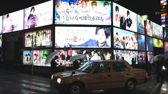 Brightly lit billboards display colorful advertisements featuring various people and text in an urban nighttime setting. A taxi is parked on the wet street while several pedestrians holding umbrellas walk by.