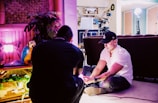 Two people are in a room engaged in a creative setting. One person is seated on the floor with lighting equipment nearby, wearing a black cap. The room includes vibrant pink lighting, a decorative parrot, plants, and an assortment of film or photography equipment.
