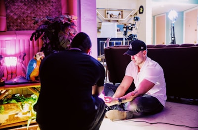 Two people are in a room engaged in a creative setting. One person is seated on the floor with lighting equipment nearby, wearing a black cap. The room includes vibrant pink lighting, a decorative parrot, plants, and an assortment of film or photography equipment.