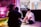 Two people are in a room engaged in a creative setting. One person is seated on the floor with lighting equipment nearby, wearing a black cap. The room includes vibrant pink lighting, a decorative parrot, plants, and an assortment of film or photography equipment.