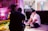Two people are in a room engaged in a creative setting. One person is seated on the floor with lighting equipment nearby, wearing a black cap. The room includes vibrant pink lighting, a decorative parrot, plants, and an assortment of film or photography equipment.
