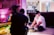 Two people are in a room engaged in a creative setting. One person is seated on the floor with lighting equipment nearby, wearing a black cap. The room includes vibrant pink lighting, a decorative parrot, plants, and an assortment of film or photography equipment.