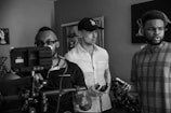 A monochrome scene depicts three individuals in a room. One person in the foreground is holding a camera rig, focused on a task, while wearing glasses and a polo shirt. Another person, wearing a cap and a button-up shirt, looks intently forward, holding a mobile device. The third individual, casually dressed in a patterned T-shirt, appears to be speaking or reacting to something, holding an object in hand. The environment contains decorative elements such as sculptures and framed art.