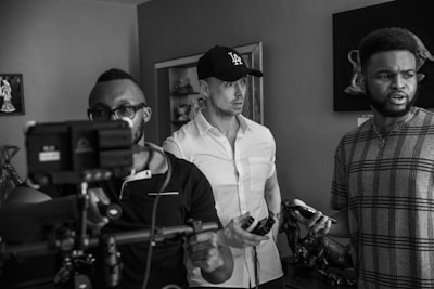 A monochrome scene depicts three individuals in a room. One person in the foreground is holding a camera rig, focused on a task, while wearing glasses and a polo shirt. Another person, wearing a cap and a button-up shirt, looks intently forward, holding a mobile device. The third individual, casually dressed in a patterned T-shirt, appears to be speaking or reacting to something, holding an object in hand. The environment contains decorative elements such as sculptures and framed art.