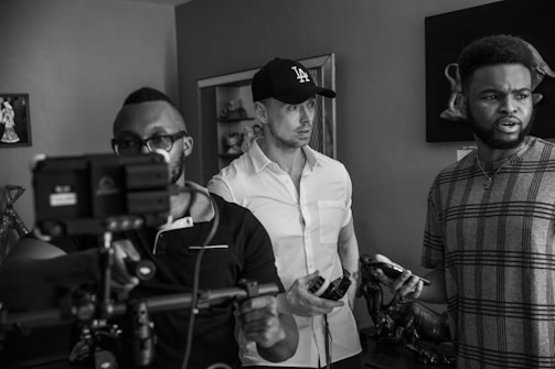 A monochrome scene depicts three individuals in a room. One person in the foreground is holding a camera rig, focused on a task, while wearing glasses and a polo shirt. Another person, wearing a cap and a button-up shirt, looks intently forward, holding a mobile device. The third individual, casually dressed in a patterned T-shirt, appears to be speaking or reacting to something, holding an object in hand. The environment contains decorative elements such as sculptures and framed art.