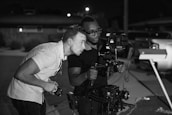 Two individuals appear to be focusing intently on a camera setup. One holds a stabilizer with a mounted camera and external monitor, indicating involvement in film or video production, possibly in an outdoor setting under low light conditions.