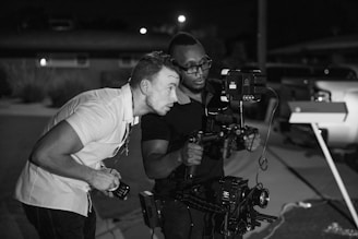 Two individuals appear to be focusing intently on a camera setup. One holds a stabilizer with a mounted camera and external monitor, indicating involvement in film or video production, possibly in an outdoor setting under low light conditions.