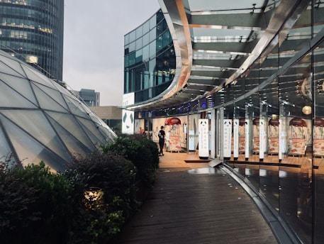 A modern architectural scene featuring a curved glass building structure reflecting light. There is a person standing near the entrance to a commercial establishment that appears to have signage with writing. To the left, a dome-shaped structure made of glass panels can be seen, surrounded by greenery.