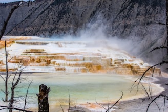 A natural limestone terrace formation with multiple cascading levels covered in steam, surrounded by rocky hills and sparse vegetation. The terraced structures exhibit various shades of white, yellow, and orange, creating a striking visual effect.
