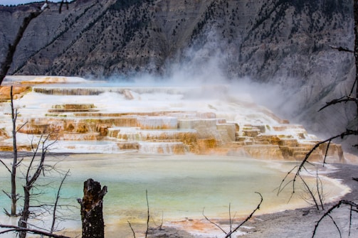 A natural limestone terrace formation with multiple cascading levels covered in steam, surrounded by rocky hills and sparse vegetation. The terraced structures exhibit various shades of white, yellow, and orange, creating a striking visual effect.