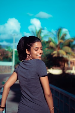 A woman with a ponytail is smiling as she stands on a bridge. The background features palm trees and a bright blue sky with scattered clouds.