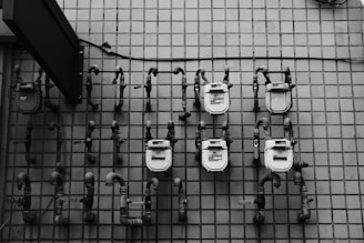 Several utility meters are mounted on a tiled wall, each connected by a series of pipes. The image is in grayscale, showcasing the industrial and geometric nature of the scene.