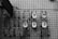 Several utility meters are mounted on a tiled wall, each connected by a series of pipes. The image is in grayscale, showcasing the industrial and geometric nature of the scene.
