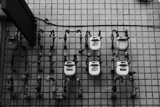 Several utility meters are mounted on a tiled wall, each connected by a series of pipes. The image is in grayscale, showcasing the industrial and geometric nature of the scene.