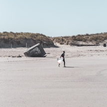 A lone surfer walks across a sandy beach carrying a surfboard, with sand dunes covered in grass in the background. A large concrete structure, possibly a bunker, is partially buried in the sand, featuring graffiti art.