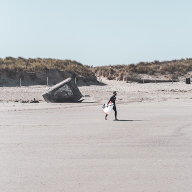 A lone surfer walks across a sandy beach carrying a surfboard, with sand dunes covered in grass in the background. A large concrete structure, possibly a bunker, is partially buried in the sand, featuring graffiti art.
