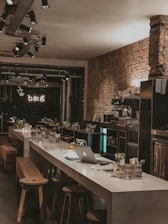 A modern, dimly lit cafe with a long white table at the center, surrounded by wooden stools. There are various items on the table, including a laptop, glasses, crockery, and a single orange. The cafe features exposed brick walls and industrial-style lighting fixtures and pipes on the ceiling. The back wall displays a neon sign reading 'b&g'.