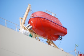 A bright orange lifeboat is securely attached to the side of a ship, hoisted on a metal frame. The background is a clear blue sky, and the ship's deck is partially visible with some safety equipment like life rings.
