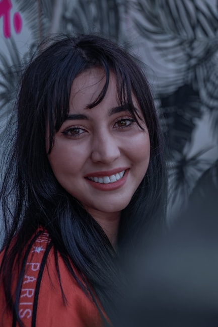 A person with long black hair and a warm smile is looking directly at the camera. The background features a blurred pattern of leaves and a hint of pink text.
