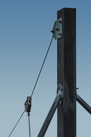 A close-up of a sturdy steel structure featuring a vertical beam with two attached metal cables. One cable is secured with a metal bracket, and the other is connected to a pulley system. The background is a clear blue sky, emphasizing the industrial design.