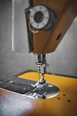 A close-up view of a sewing machine needle and presser foot, showcasing the intricate metal parts and threading mechanism against a textured background. The focus is on the metallic components, highlighting the precision and craftsmanship involved in sewing.