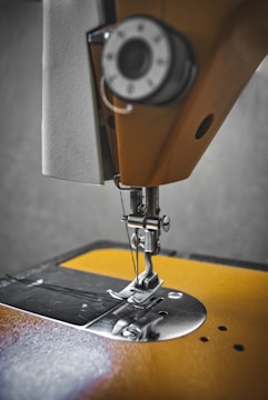 A close-up view of a sewing machine needle and presser foot, showcasing the intricate metal parts and threading mechanism against a textured background. The focus is on the metallic components, highlighting the precision and craftsmanship involved in sewing.